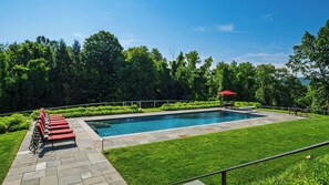 Ein rechteckiger Pool mit einer umlaufenden Pflasterterrasse, einem roten Sonnenschirm und einer Reihe roter Liegestühle.