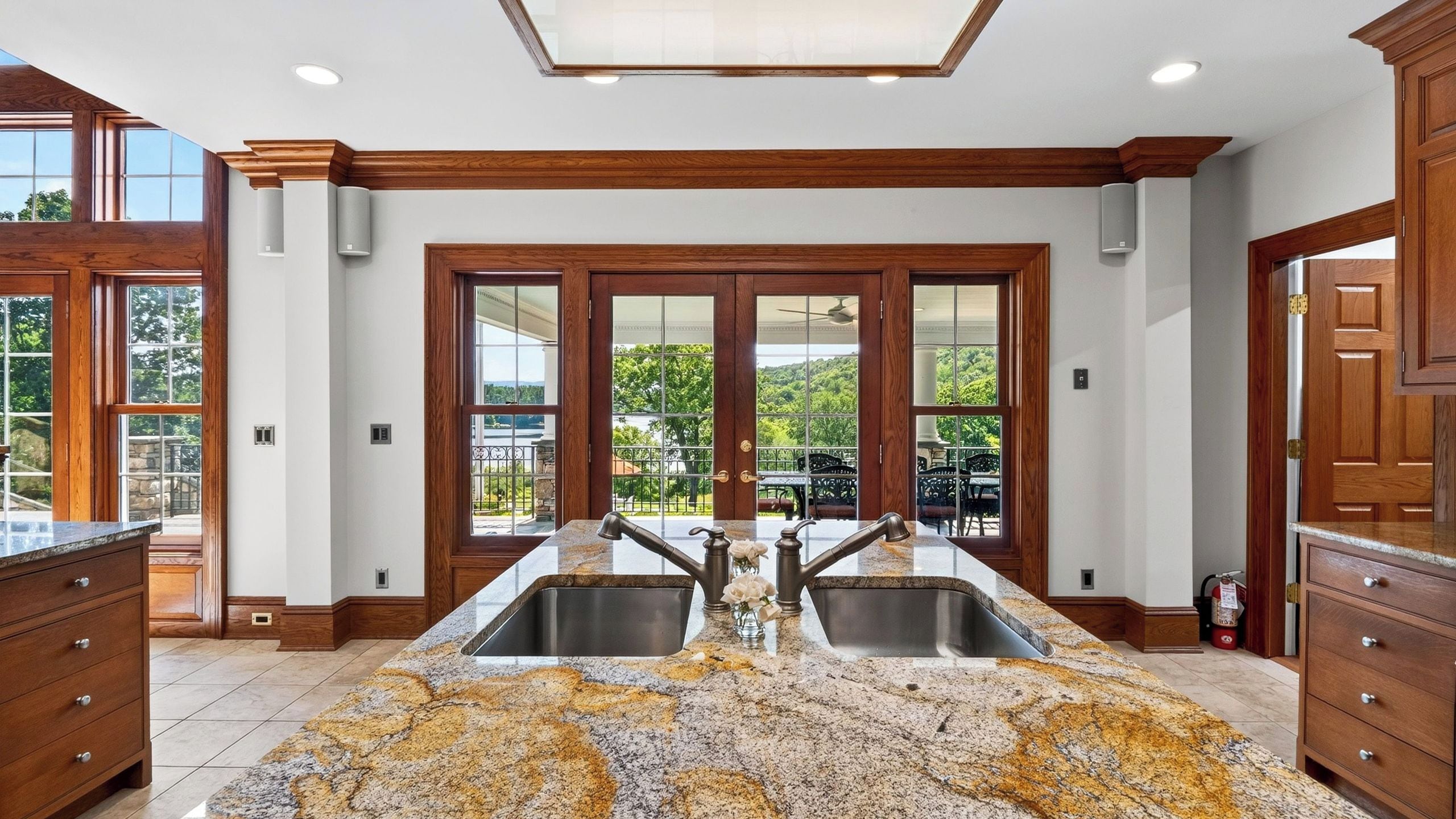 A kitchen with a central island, double sinks, wooden cabinetry, and large windows offering a view of greenery.