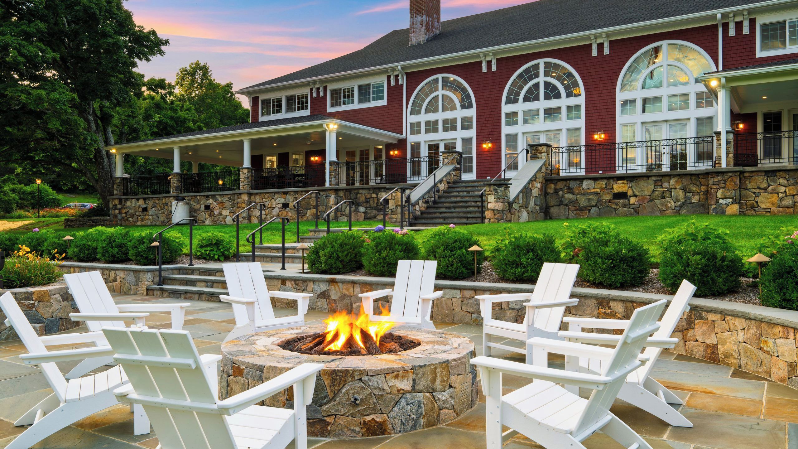 A stone fire pit surrounded by white chairs, set on a patio with a stone wall and steps leading to a covered porch.