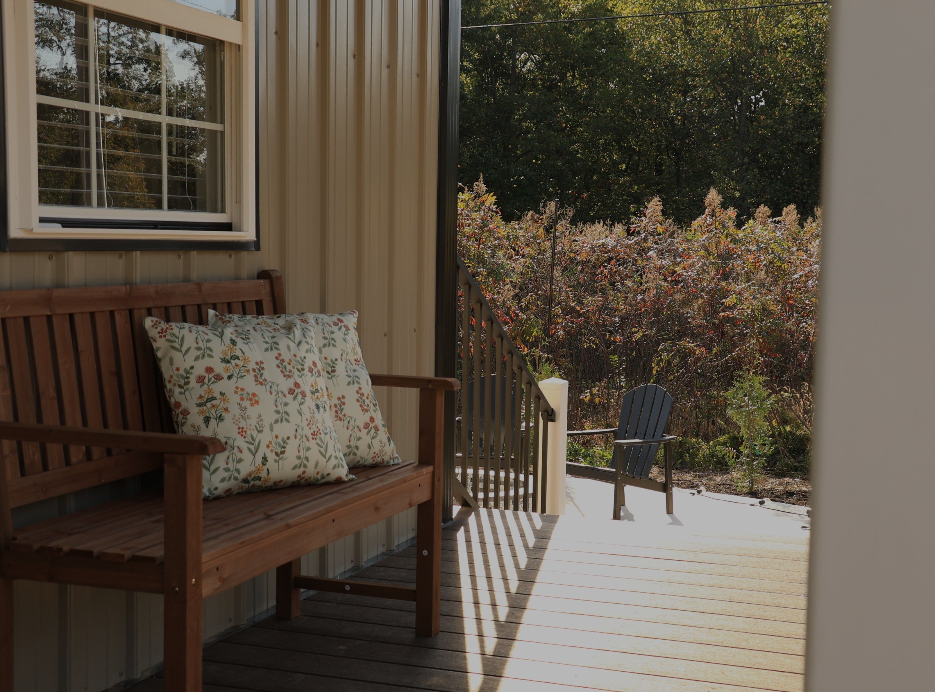 A wooden bench with a floral cushion, a chair, and a view of a garden with autumn foliage.