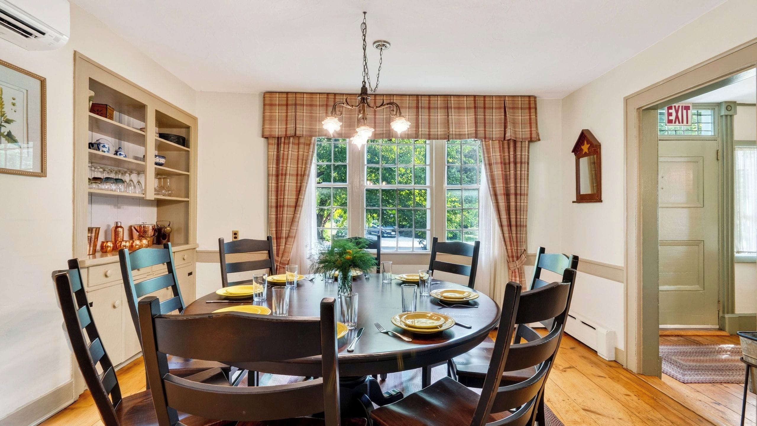 A dining room with a round table, six chairs, a chandelier, and a view of greenery outside.
