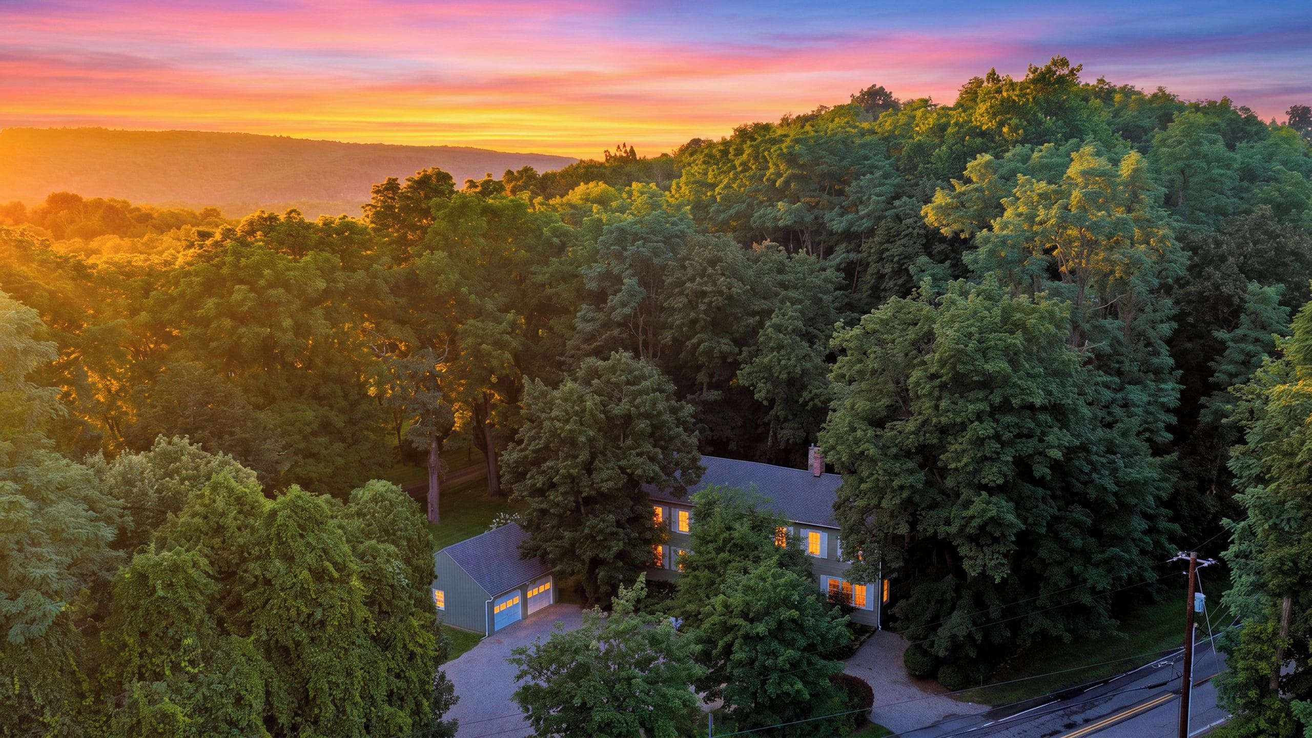 A residential area with a house surrounded by dense trees, a clear sky, and a distant mountain range.