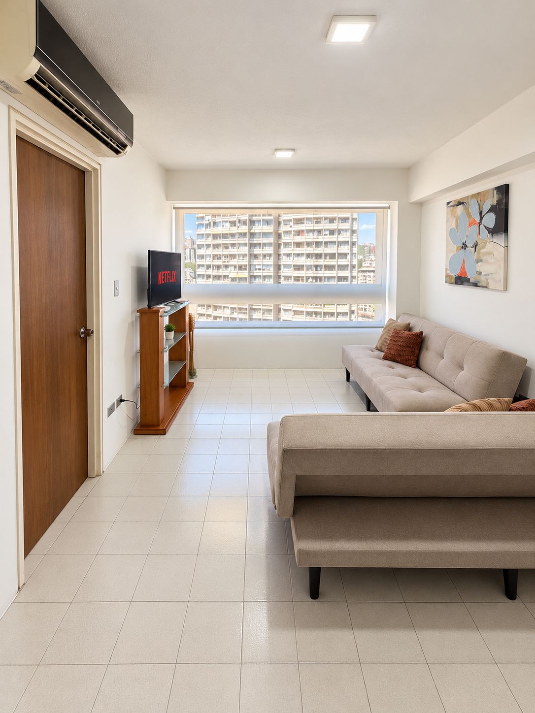 A living room with a sofa, a TV on a stand, a window with a city view, and a door.