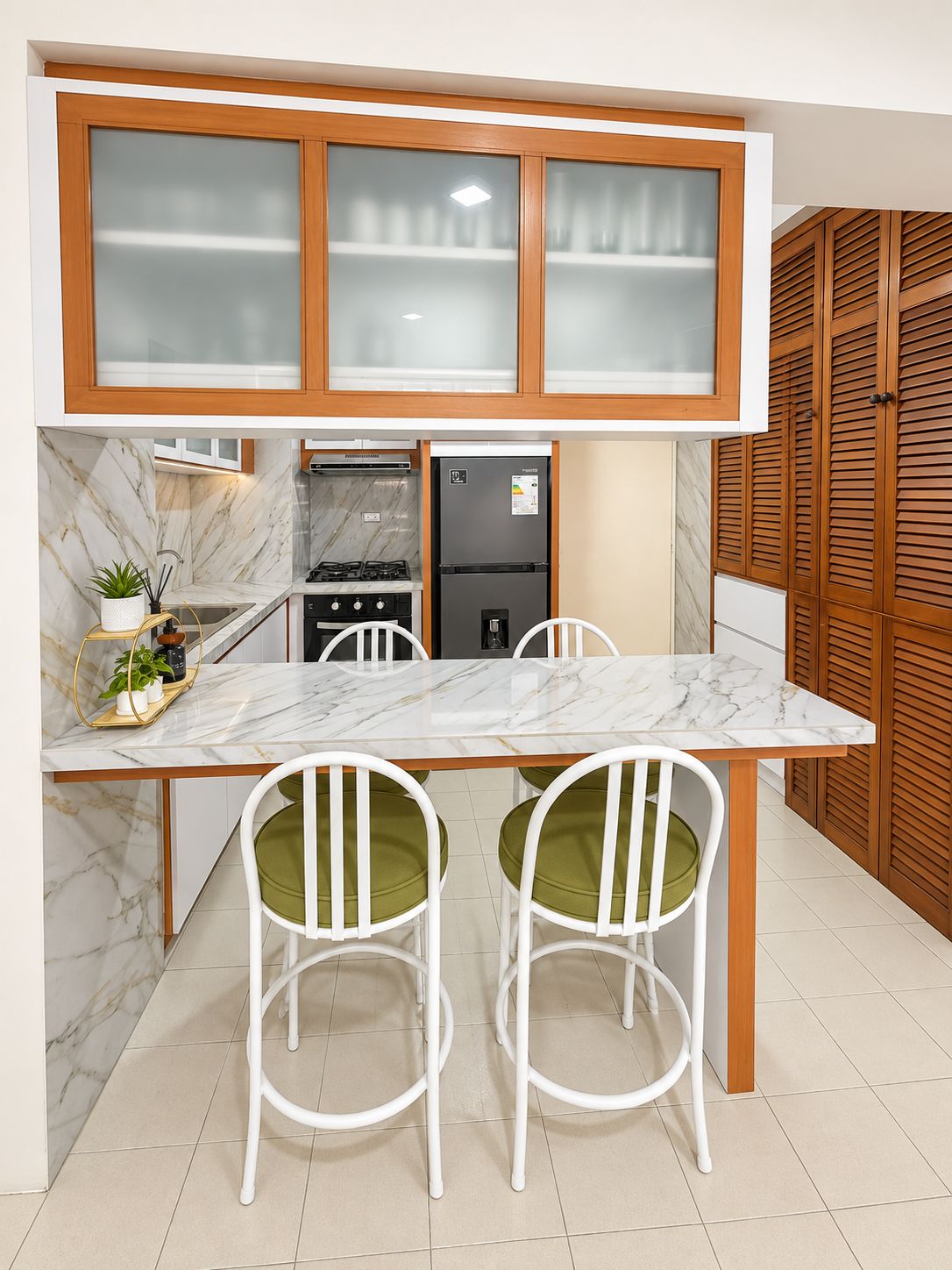 A compact kitchen with a marble countertop, two white chairs with green cushions, a black refrigerator, and wooden cabinets with frosted glass doors.