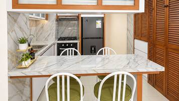 A compact kitchen with a marble countertop, two white chairs with green cushions, a black refrigerator, and wooden cabinets with frosted glass doors.