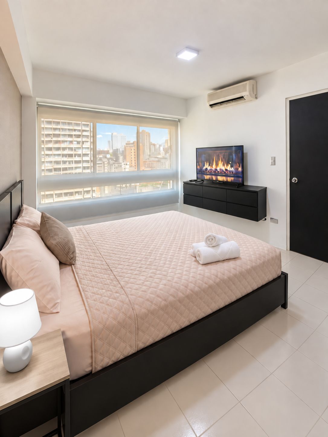 Une chambre moderne avec un grand lit, un téléviseur posé sur un meuble noir et une vue sur la ville par la fenêtre.