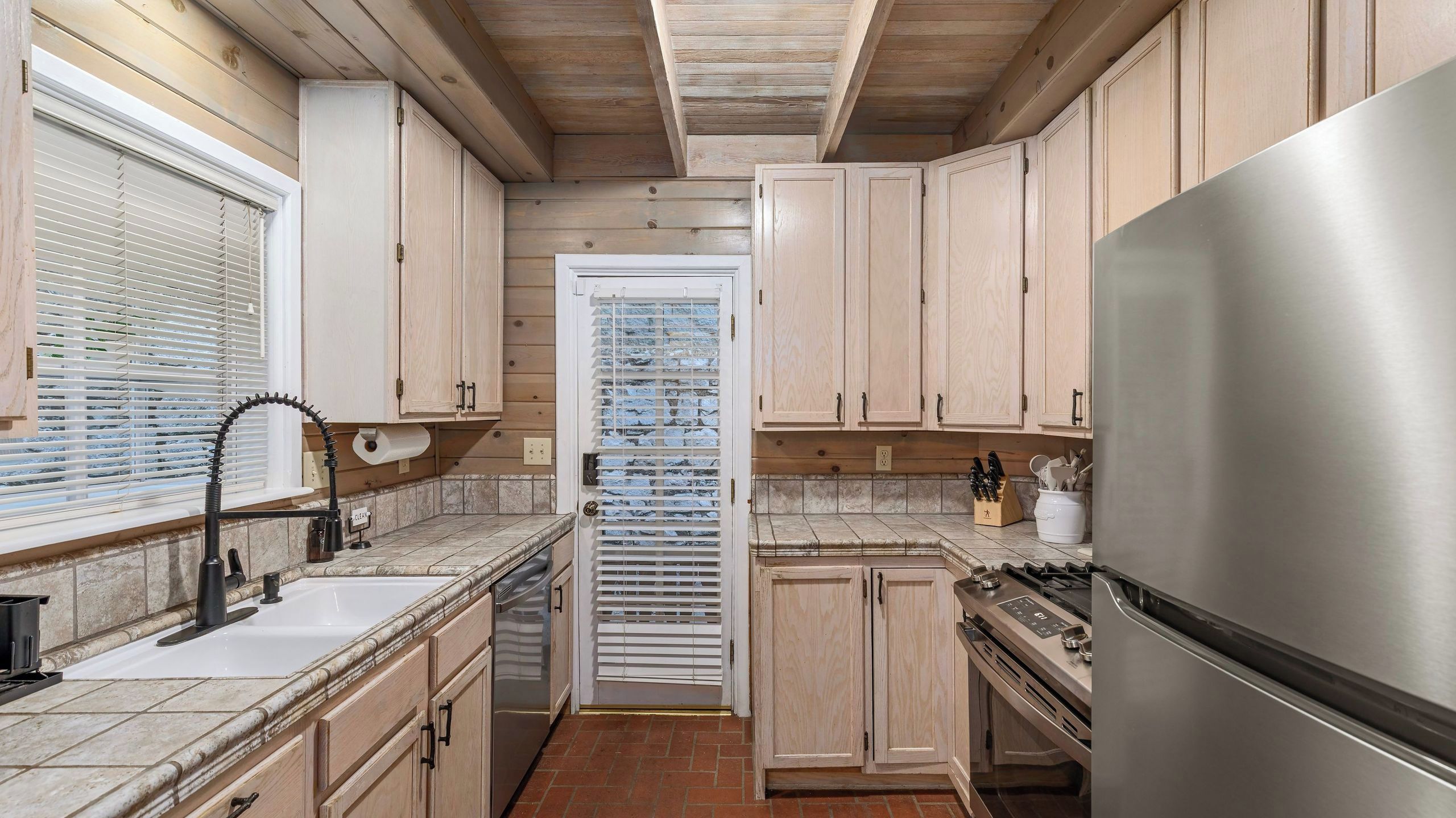 A kitchen with wooden cabinets, a stainless steel refrigerator, a sink, and a window with blinds.