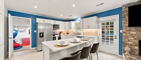 A modern kitchen with white cabinets, a central island with bar stools, and a stone fireplace.