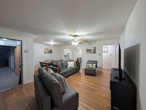 Ein modernes Wohnzimmer mit einer Couch, einem Sessel und einem Fernseher. Es gibt einen Essbereich mit Tisch und Stühlen, und im Hintergrund eine Küche.