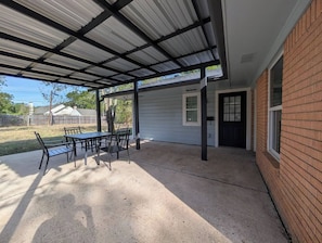 Ein überdachter Patio-Bereich mit Tisch und Stühlen, eine Ziegelwand und eine Tür zu einem Haus.