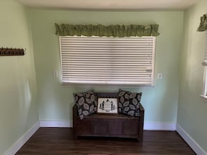 Una habitación con ventana, un banco con almohadones decorativos y un estante en la pared.