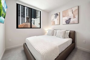Een slaapkamer met een groot bed, een raam met uitzicht op de stad en twee abstracte schilderijen aan de muur.