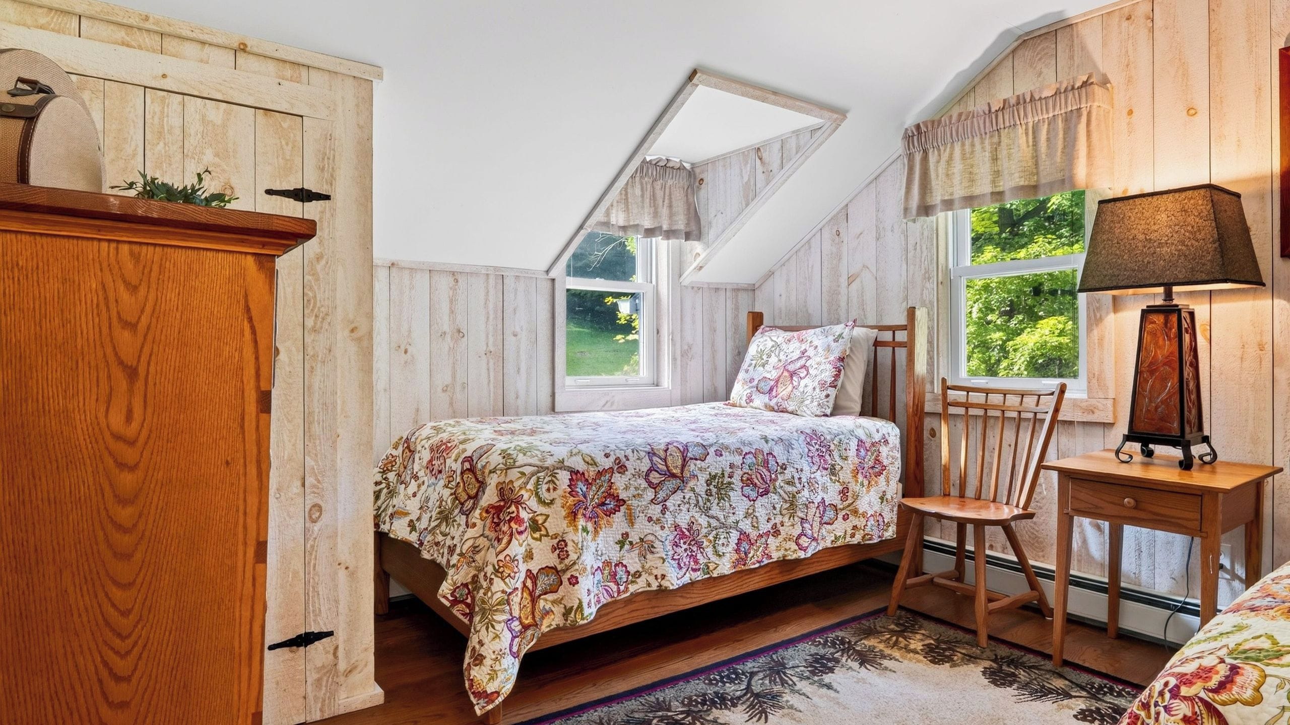 A bedroom with a sloped ceiling, a bed with floral bedding, a wooden chair, a nightstand, and a lamp.