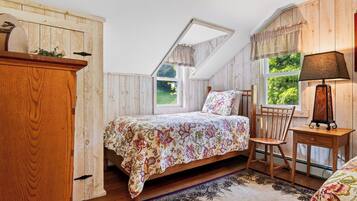 A bedroom with a sloped ceiling, a bed with floral bedding, a wooden chair, a nightstand, and a lamp.