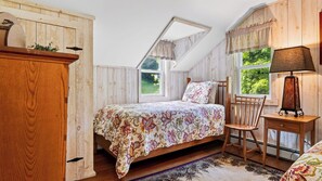 Quarto com teto inclinado, cama com roupa de cama florida, cadeira de madeira, mesa de cabeceira e abajur.