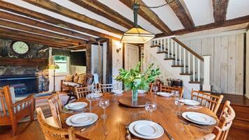 A dining room with a wooden table set for six, surrounded by wooden chairs. A chandelier hangs from the ceiling, and a fireplace is visible in the background.