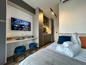 A modern hotel room with a bed, a TV mounted on the wall, a small kitchenette, and a seating area with stools. - Setin_2302 · Studio no 23º andar | 1 min do Metrô República (Santa Ifigênia)