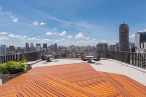 A rooftop terrace with a wooden deck, outdoor seating, and a city skyline view. - Setin_2302 · Studio no 23º andar | 1 min do Metrô República (Santa Ifigênia)