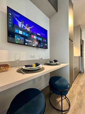 A modern kitchen with a flat-screen TV mounted above a counter, two bar stools, and a dining area with a basket and glasses. - Setin_2302 · Studio no 23º andar | 1 min do Metrô República (Santa Ifigênia)