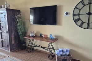 A room with a television mounted on the wall, a decorative clock, a wooden table with a fish sculpture, and a basket with towels. - Elevated Bella Sirena Views: 2nd Floor Luxury (Puerto Peñasco)