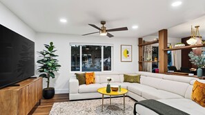 Un salon moderne avec un canapé d’angle, un téléviseur à écran plat, un ventilateur de plafond et une grande fenêtre.
