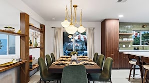 A dining area with a wooden table set for six, surrounded by green upholstered chairs, a chandelier, and a view of a tropical scene through a window. - The Royal Palms by Avantstay Pool + Hot Tub + Game Room + Fire Pit (Palm Springs)