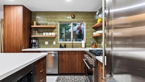 A modern kitchen with wooden cabinets, a built-in oven, and a window with a view of palm trees. - The Royal Palms by Avantstay Pool + Hot Tub + Game Room + Fire Pit (Palm Springs)
