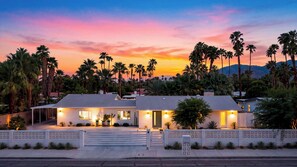 A two-story house with a covered patio and a gated entrance, surrounded by palm trees and a mountain backdrop. - The Royal Palms by Avantstay Pool + Hot Tub + Game Room + Fire Pit (Palm Springs)