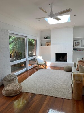 A modern living room with a fireplace, a ceiling fan, and a large window overlooking a garden. - Avalon home flat 5 minute stroll to village (Avalon Beach)