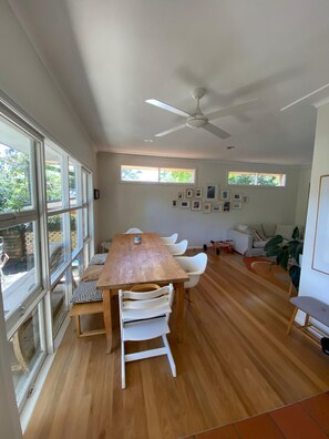 A spacious room with a wooden dining table, white chairs, a ceiling fan, and large windows. - Avalon home flat 5 minute stroll to village (Avalon Beach)
