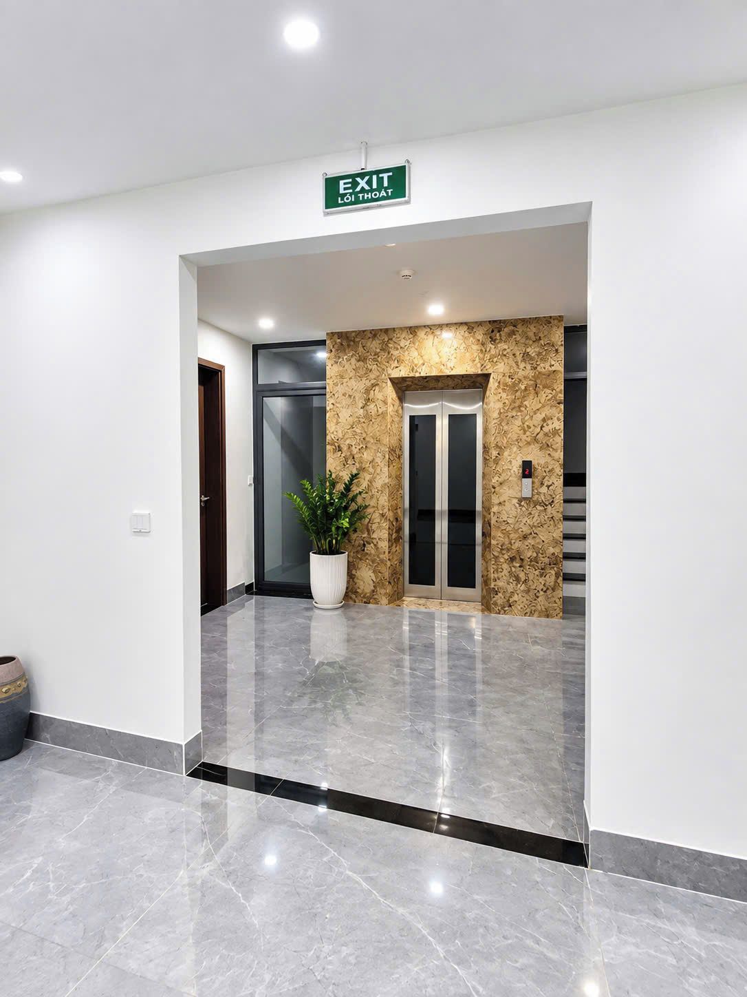 A modern lobby with a marble floor, an exit sign, a potted plant, and an elevator.