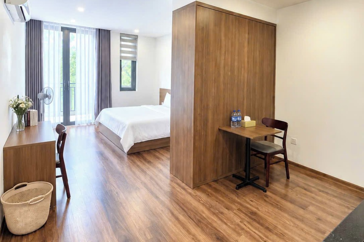 A modern hotel room with a wooden floor, a bed, a desk with a chair, a small table with bottles, and a wall-mounted air conditioner.