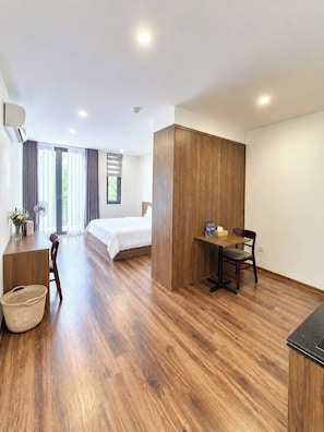 A modern hotel room with a wooden floor, a bed, a desk with a chair, a small table with bottles, and a wall-mounted air conditioner. - Nam Hai Hotel & Apartment (Vĩnh Phúc)