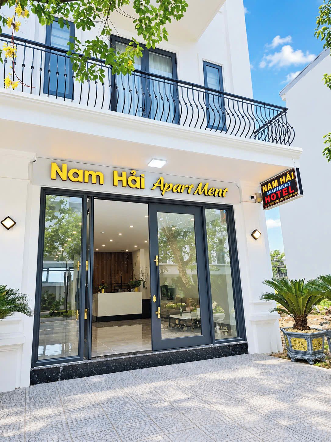 Een hotelentree met een glazen gevel, een receptiebalie en een bordje met de tekst 'Nam Hai Apartment Hotel'.