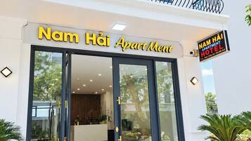 Een hotelentree met een glazen gevel, een receptiebalie en een bordje met de tekst 'Nam Hai Apartment Hotel'.