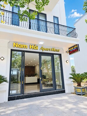 Entrada de un hotel con fachada de vidrio, una recepción y un cartel que indica 'Nam Hai Apartment Hotel'.