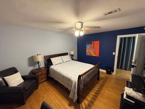 A bedroom with a bed, a nightstand, a lamp, a ceiling fan, a painting on the wall, and a door leading to another room. - Terrific 3-bedroom building, fully furnished with AC and Heat in Kansas City. (Kansas City)