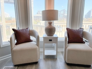 Una habitación con dos sillones, una mesita auxiliar con una lámpara y vistas a edificios a través de amplias ventanas.