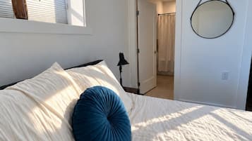 Un dormitorio con cama, una ventana con planta, una puerta que da a otra habitación y un espejo montado en la pared.