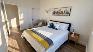 Un dormitorio con cama, una silla, una mesita de noche, una lámpara y un cuadro con una vista urbana en la pared.