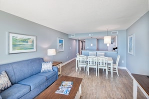 A living room with a blue sofa, a coffee table with magazines, a dining area with white chairs, and a kitchen in the background. - Stunning View Amazing Oceanfront on the 12th Floor (North Myrtle Beach)