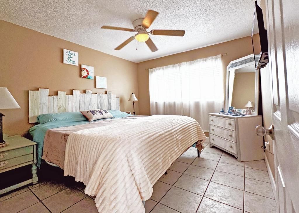 Un dormitorio con cama, mesita de noche, cómoda, ventilador de techo y ventana con cortinas.