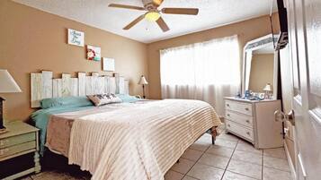Un dormitorio con cama, mesita de noche, cómoda, ventilador de techo y ventana con cortinas.