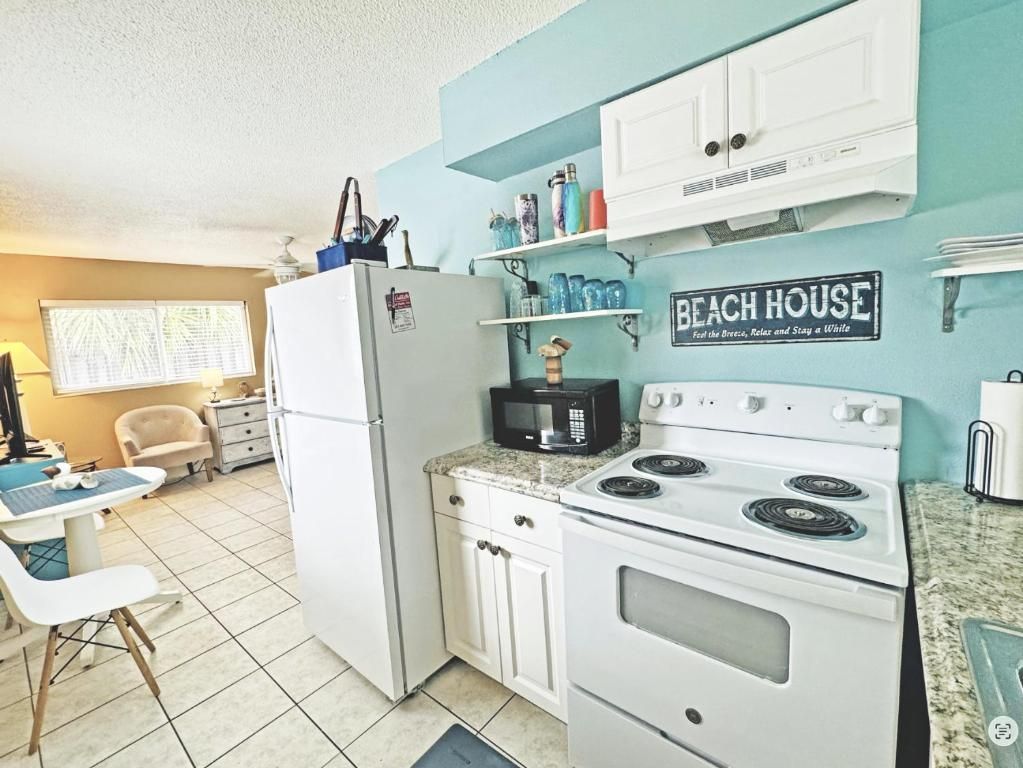 A compact kitchen with a white refrigerator, stove, and microwave. There is a sign on the wall that reads 'BEACH HOUSE.'