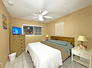 A bedroom with a bed, a nightstand, a TV, a ceiling fan, and a window with blinds.