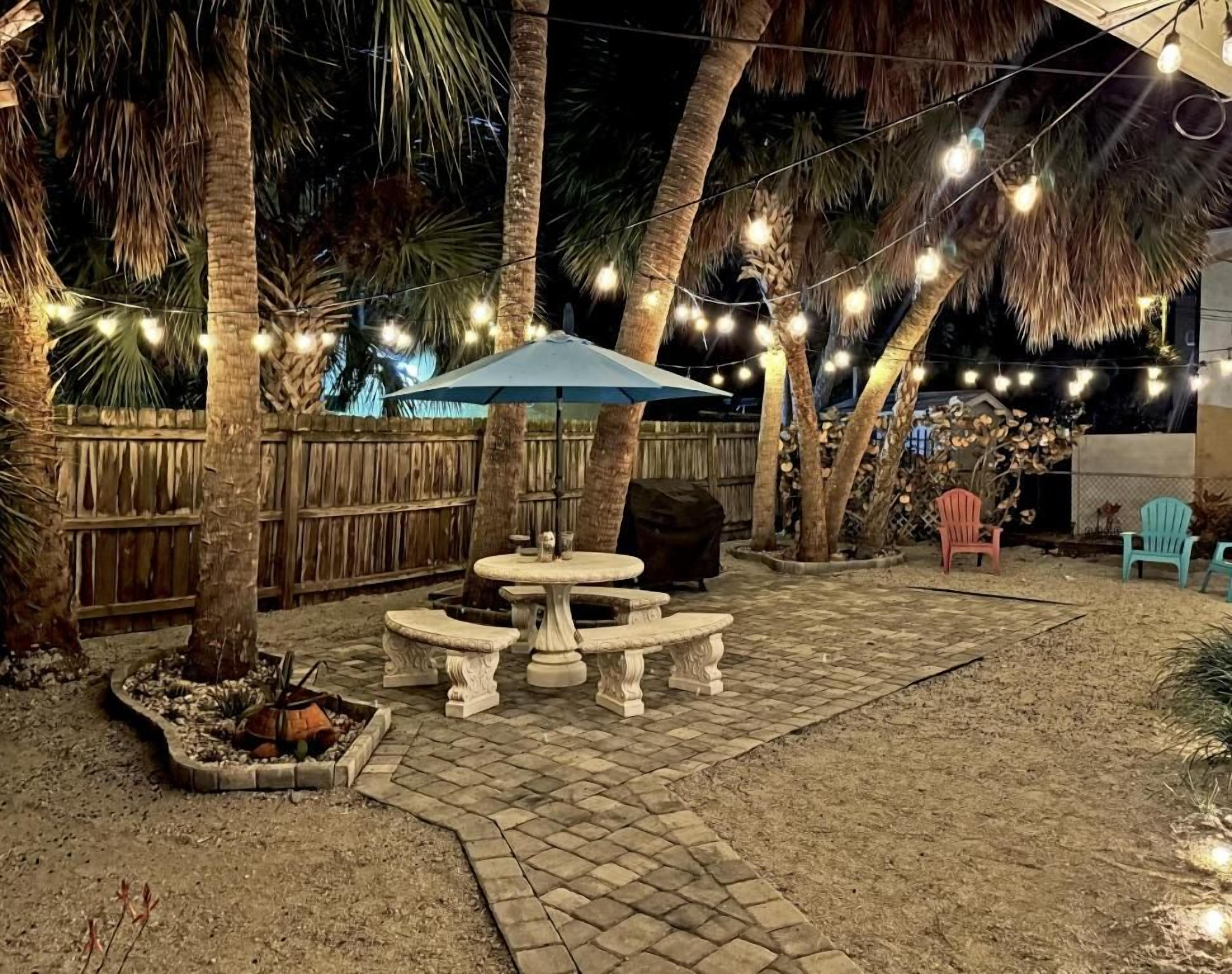 An outdoor patio area with a stone table and benches, surrounded by palm trees and string lights.