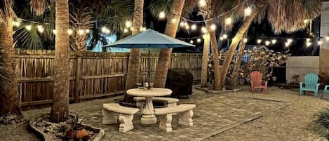 An outdoor patio area with a stone table and benches, surrounded by palm trees and string lights.