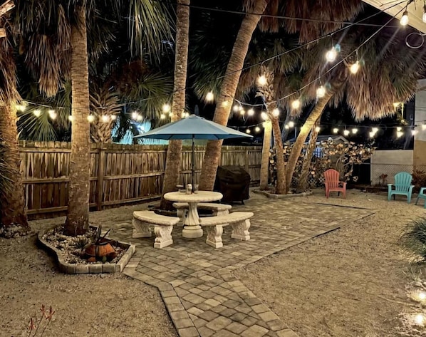 An outdoor patio area with a stone table and benches, surrounded by palm trees and string lights.