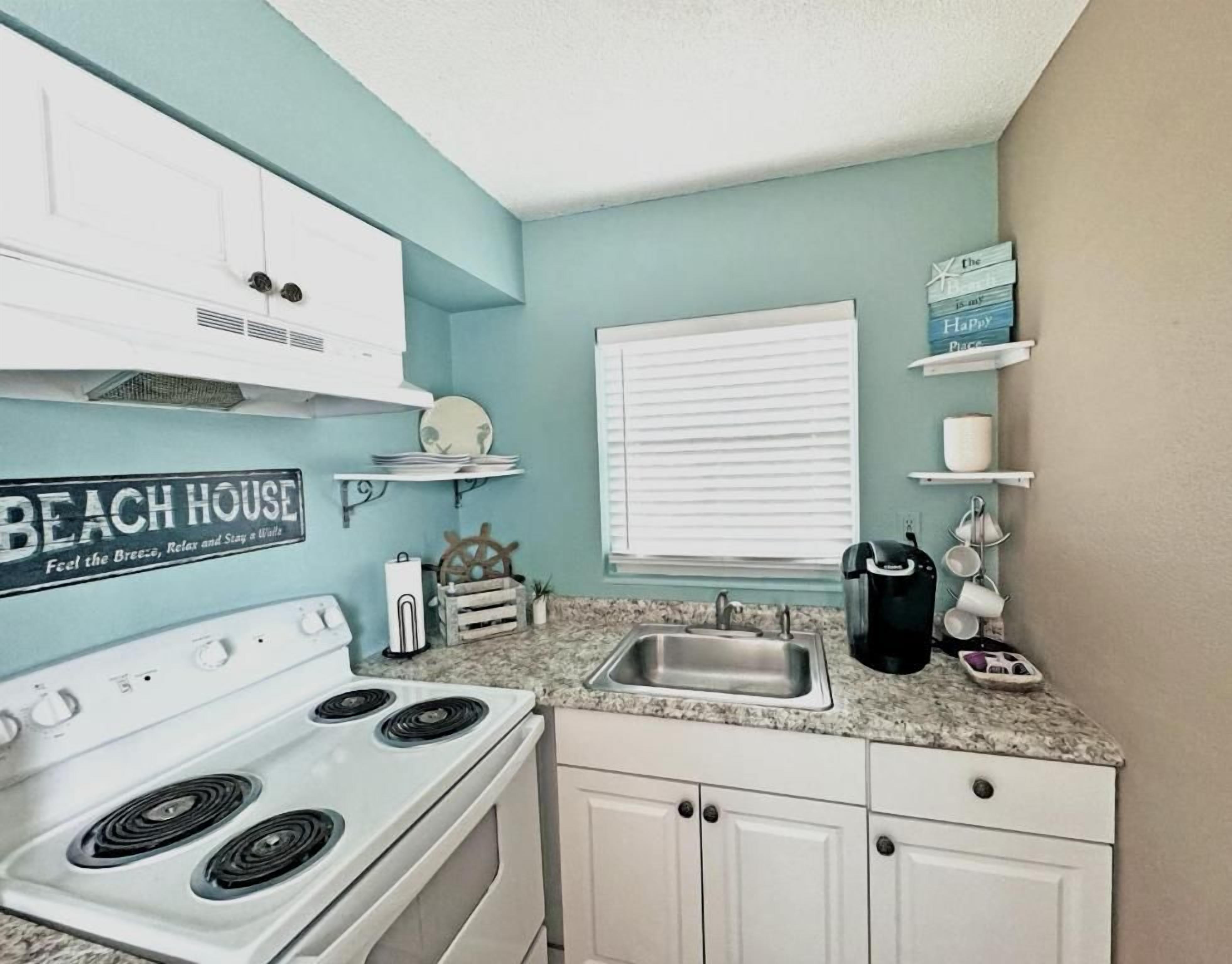 A kitchen with a white stove, a sink, and a window with blinds. There is a sign on the wall that reads 'BEACH HOUSE.'