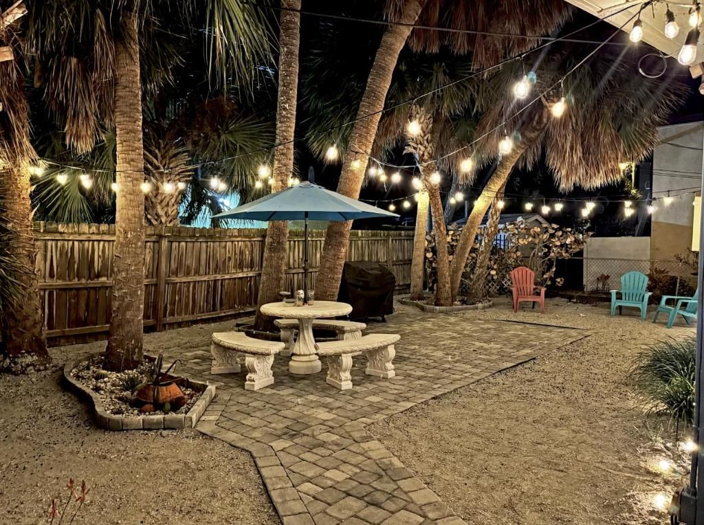 An outdoor patio area with a stone table and benches, surrounded by palm trees and string lights.